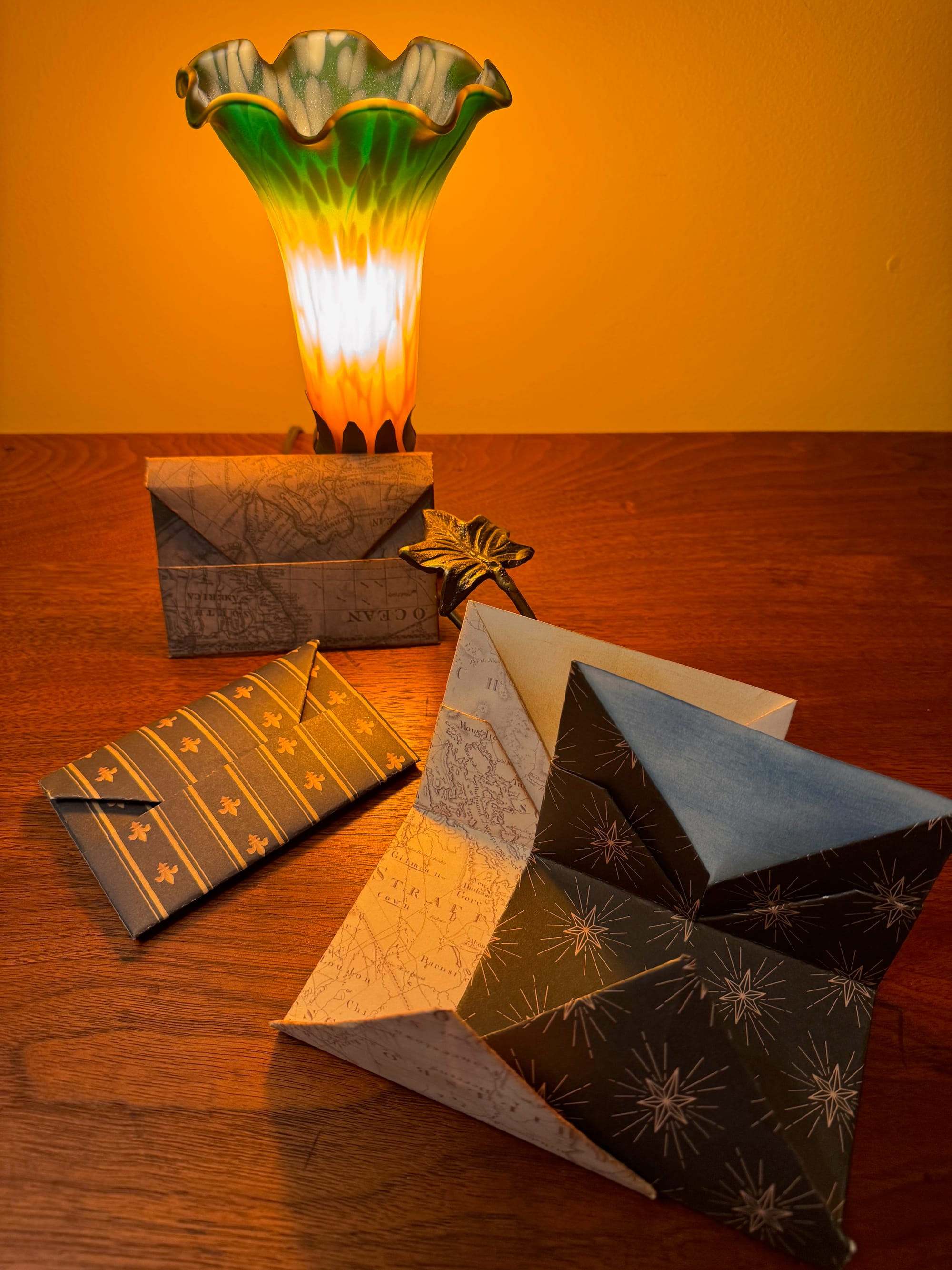 Several unusually folded envelopes made of patterned paper lay on a rich wood surface lit by a lamp that looks like a glass flower.