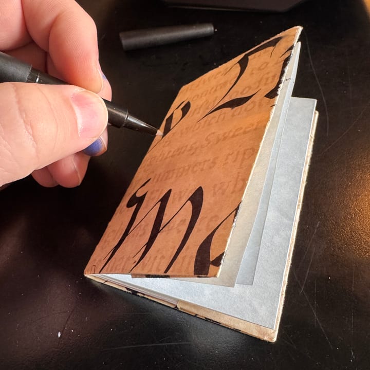 a small folded notebook with a calligraphic cover with a hand holding a felt-tip pen over it.