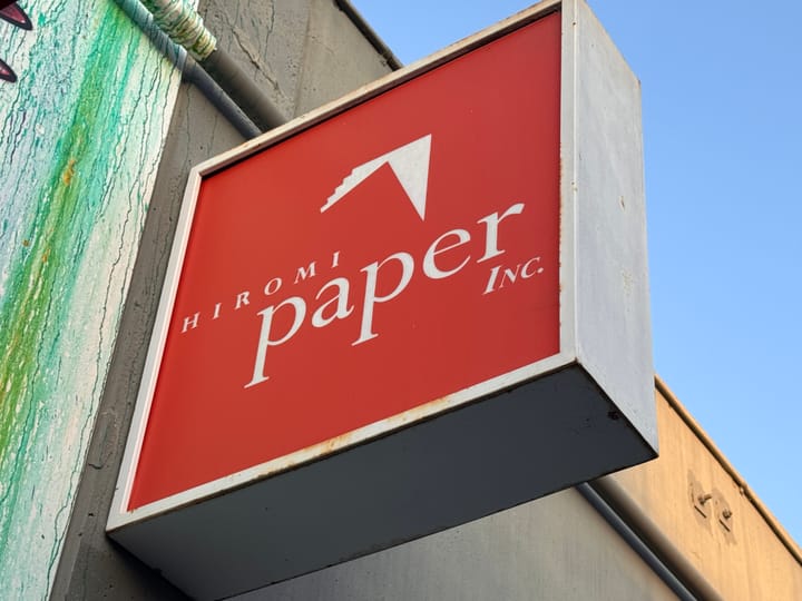 A sign sticking out from an industrial-looking wall that reads "Hiromi Paper Inc" with an icon resembling an origami bird.